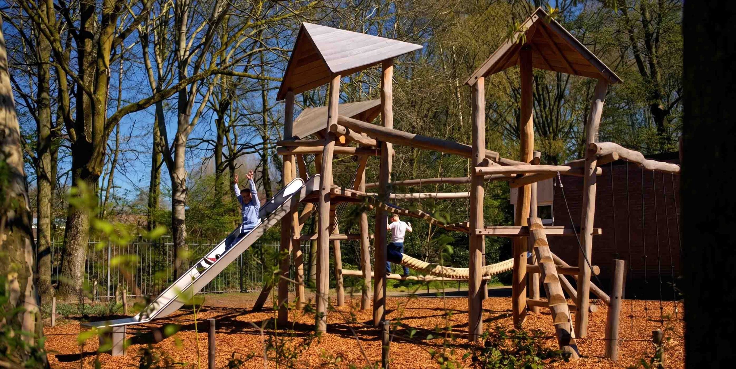 Houten speeltoestel met glijbaan en klimtouw op speelplaats tussen bomen, kinderen spelen erop.