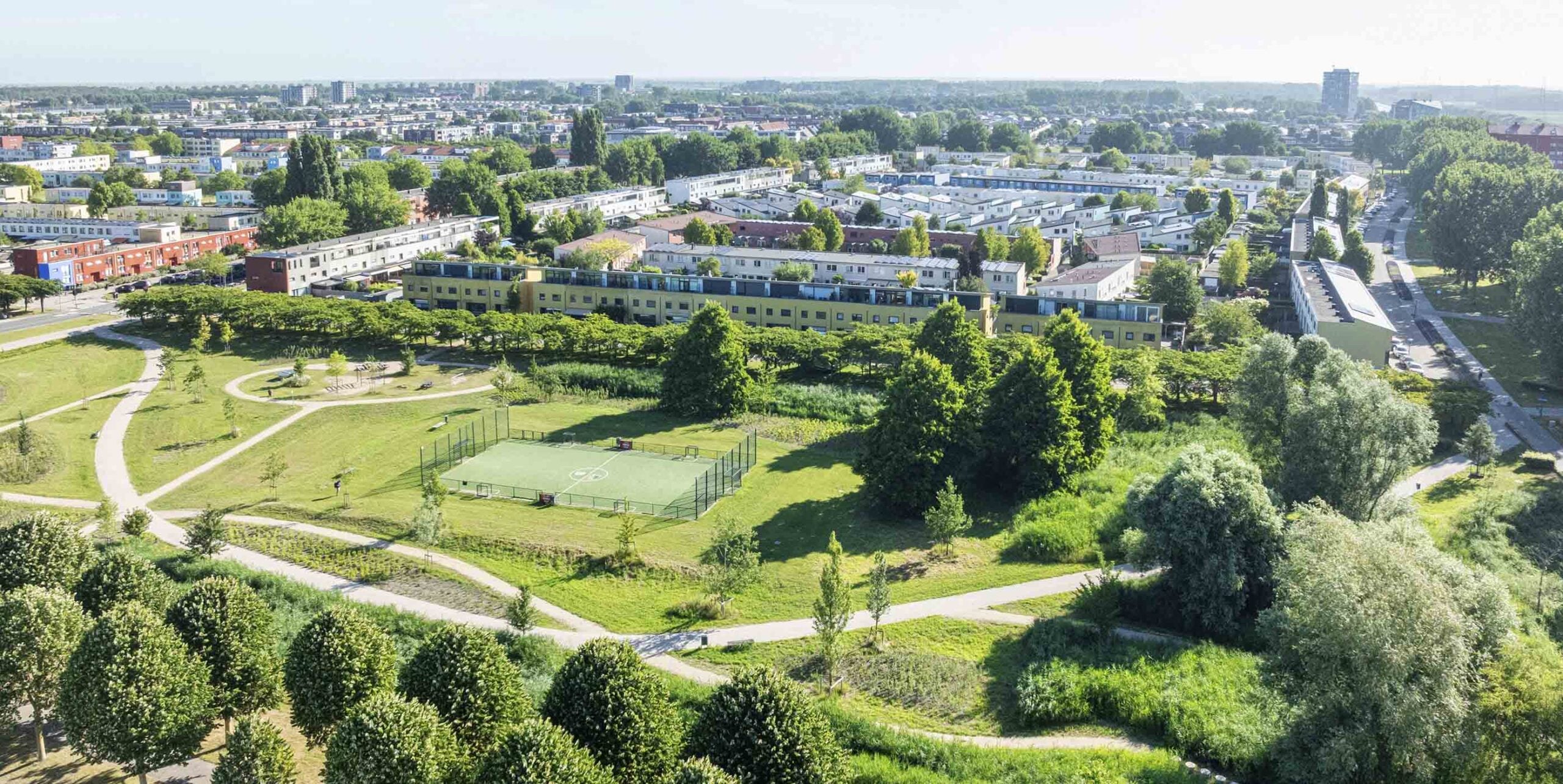 Luchtfoto van een groen stadspark met wandelpaden, trapveld en speeltoestellen tussen woonwijken.