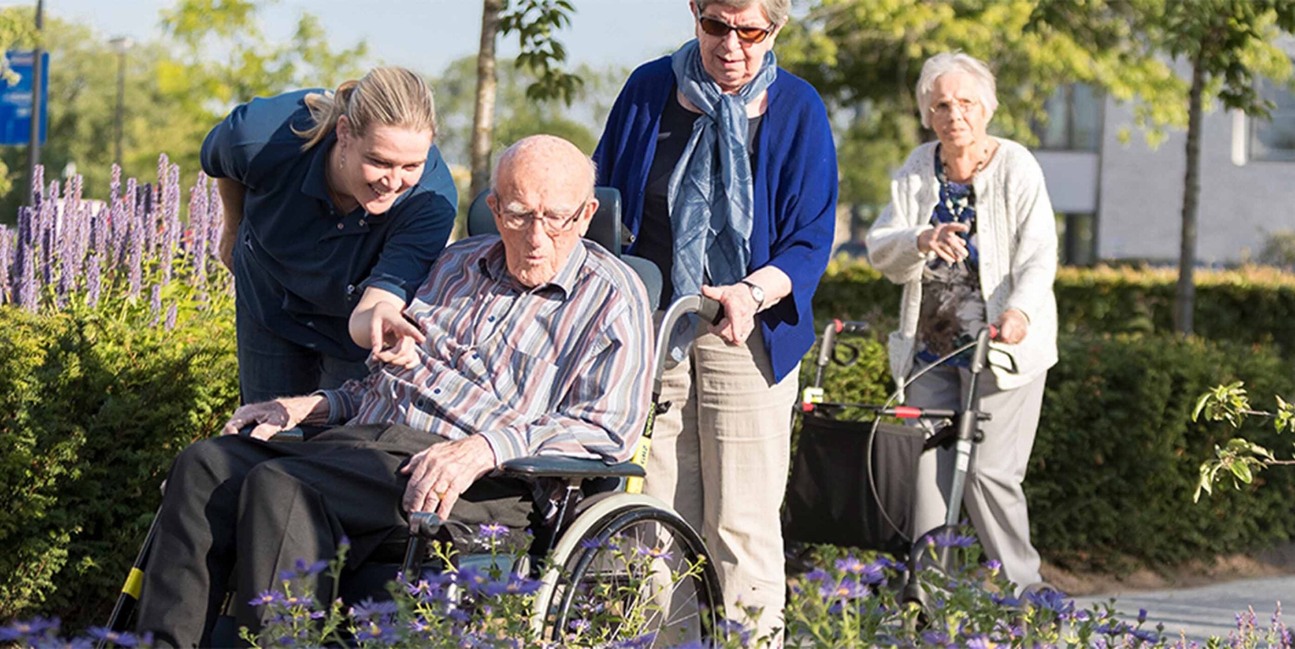 Bewoners van een zorglocatie genieten met begeleiding van een groene tuin met bloeiende border.