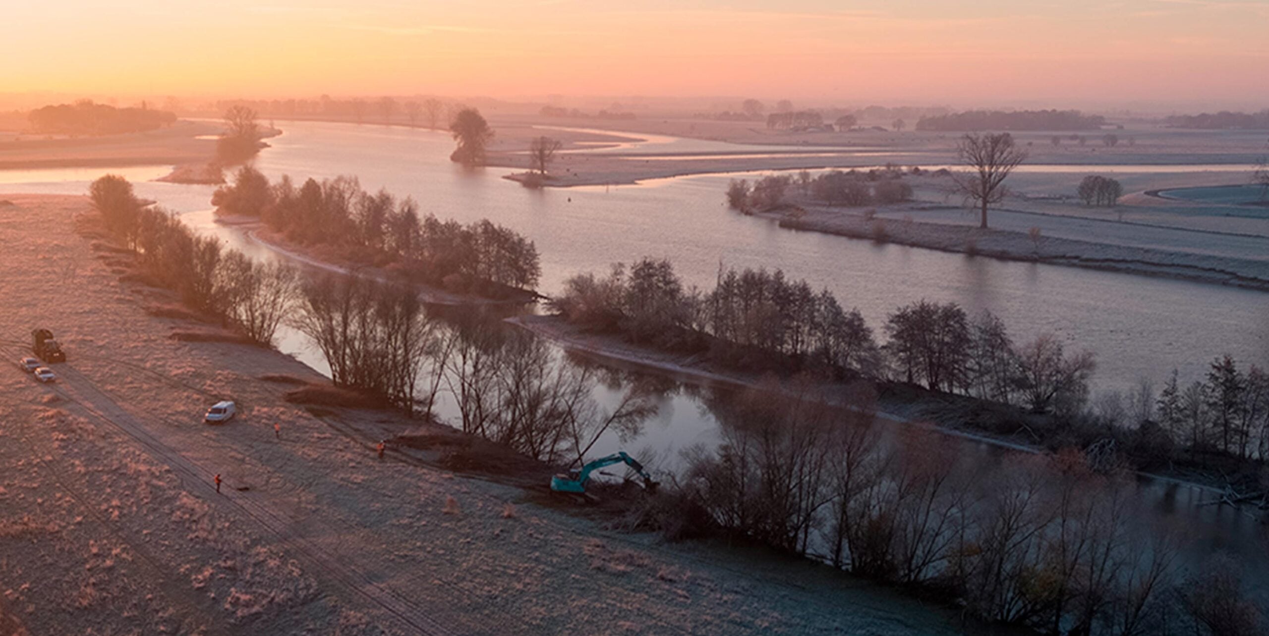 Luchtfoto van medewerkers en een graafmachine die bij zonsopkomst oeverwerk uitvoeren langs een rivier.
