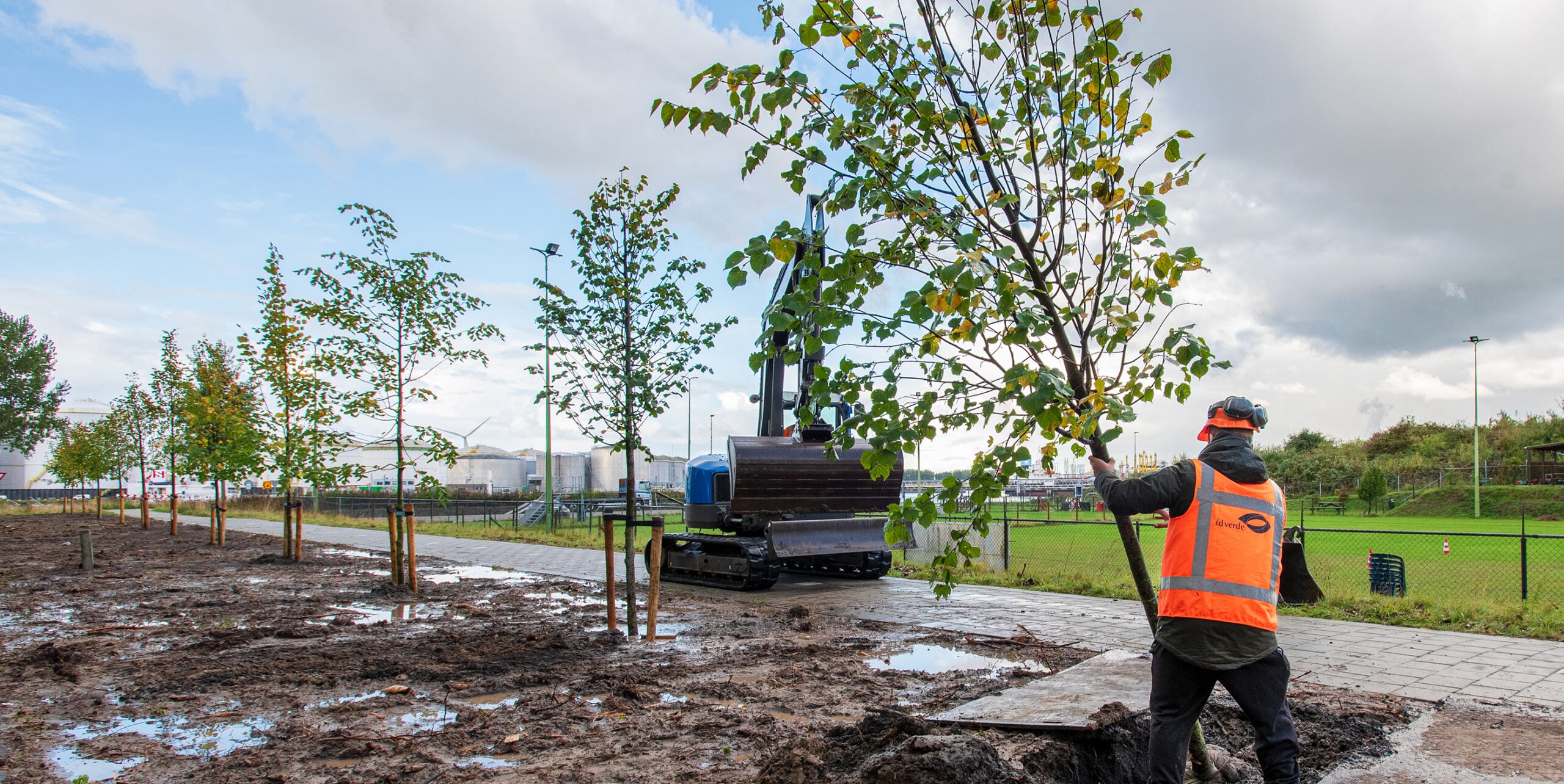 Medewerker van idverde plant een boom langs een pad in een industriegebied, met een graafmachine op de achtergrond.
