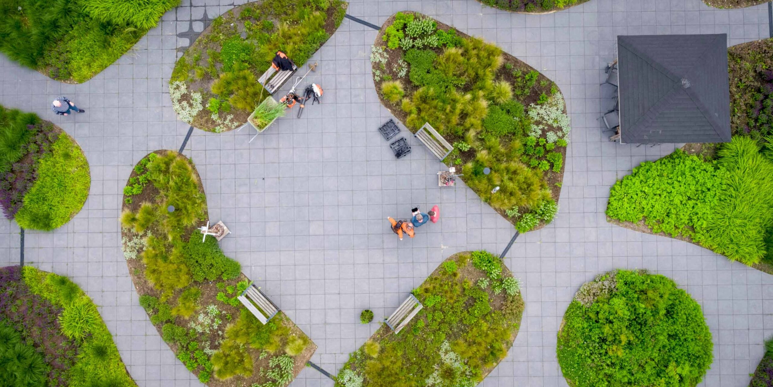 Luchtfoto van medewerkers die groenperken onderhouden op een stedelijk plein met plantsoenen tussen betegelde paden.