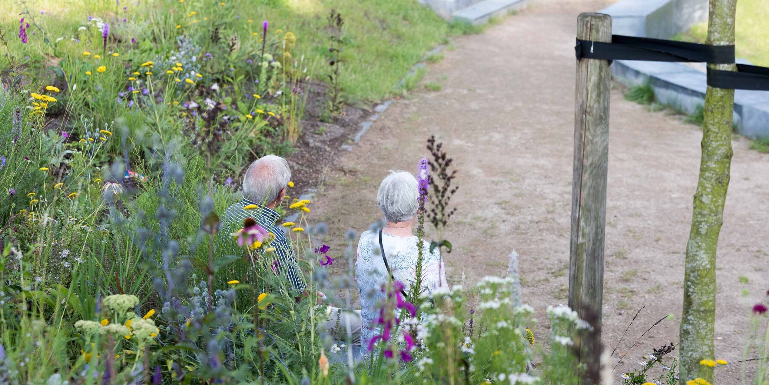 Ouder echtpaar loopt over een pad langs een bloemrijke border met vaste planten op een begraafplaats.