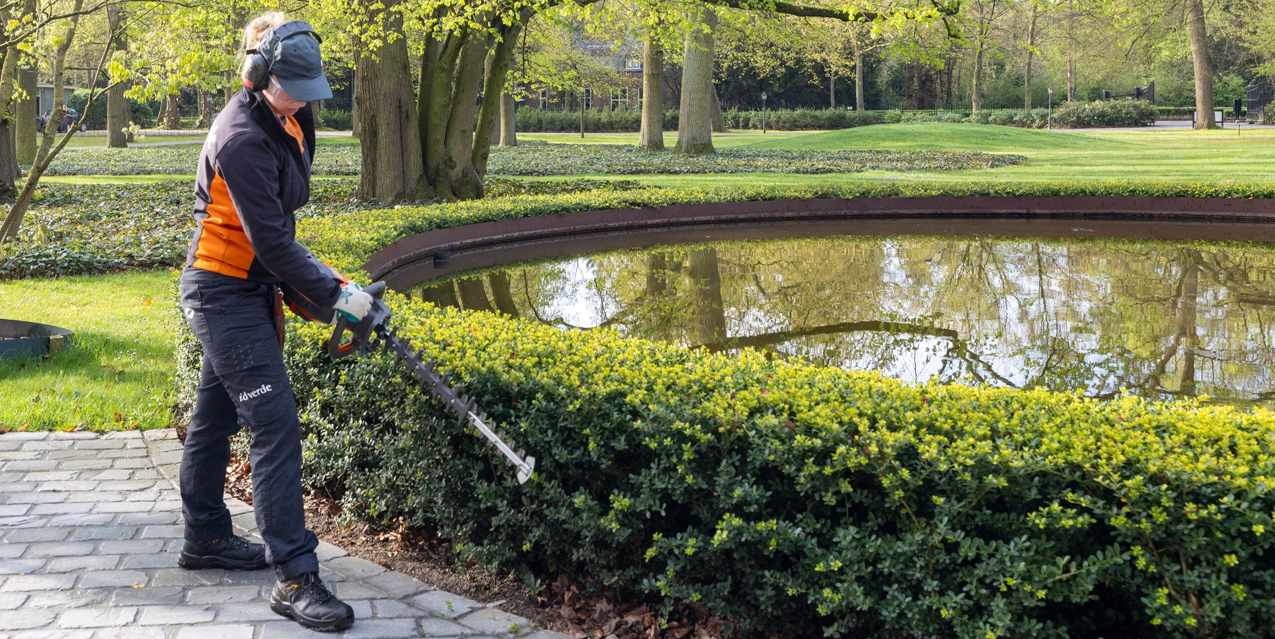 Medewerker in idverde-uniform snoeit een heg langs een vijver in een park.