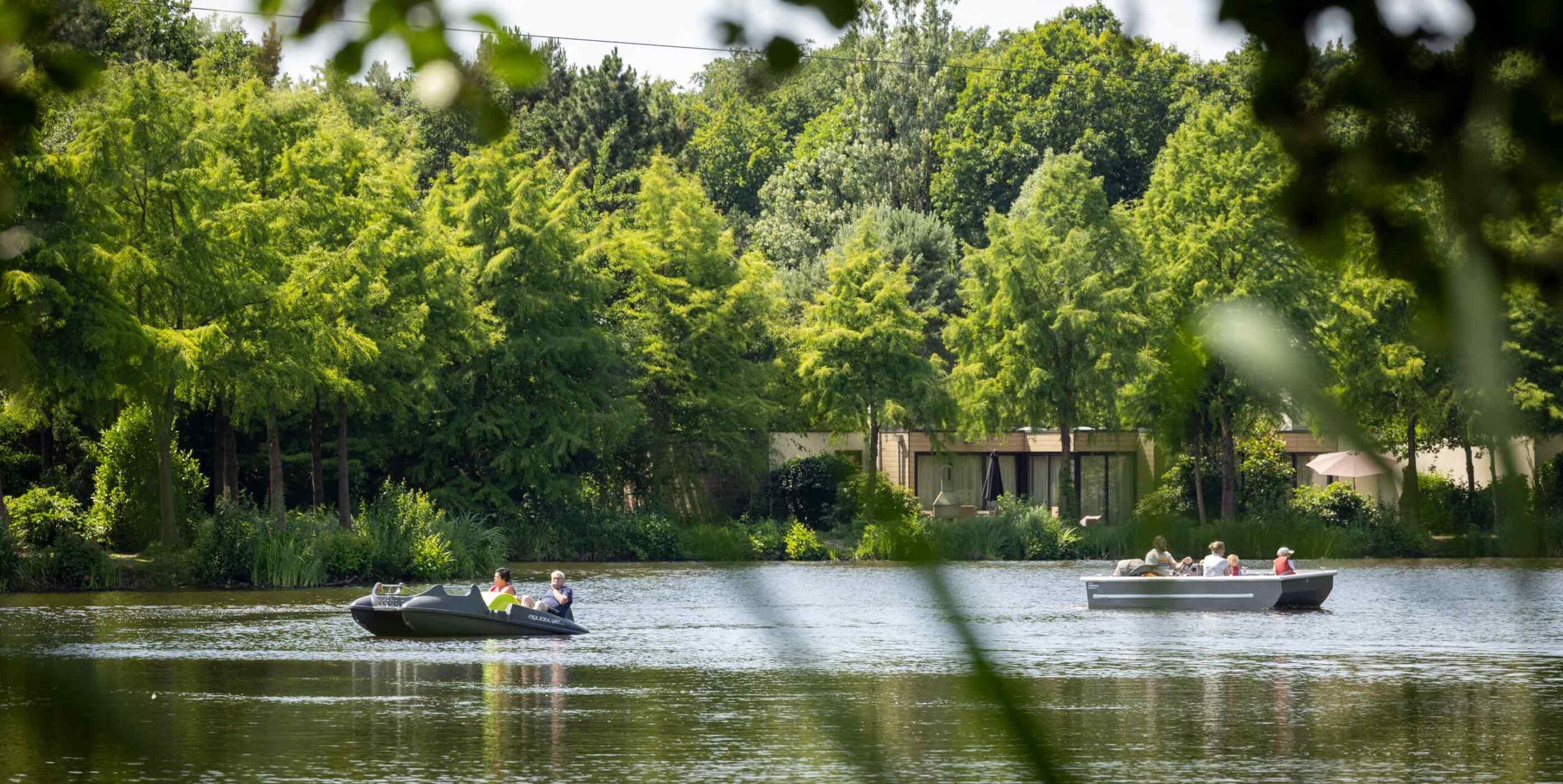 Vakantiepark met bungalows omringd door groen aanplant aan een meer waar bootjes varen.
