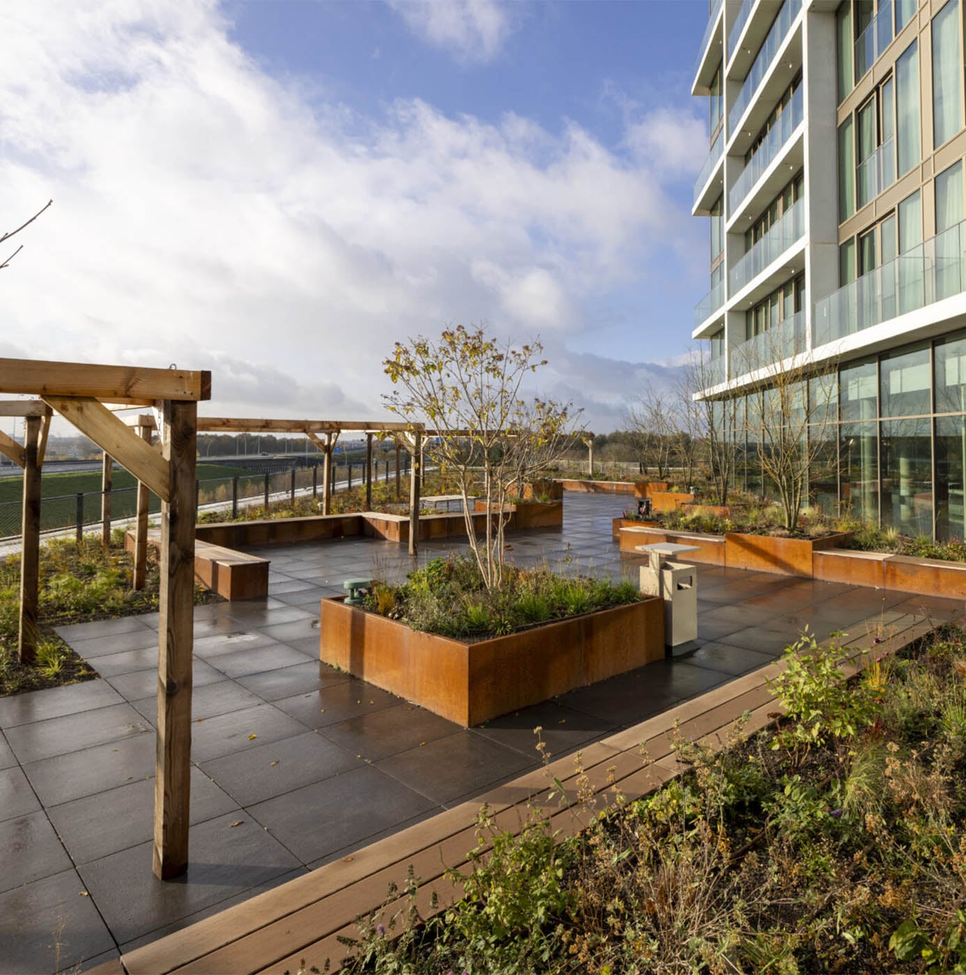 Daktuin met cortenstalen plantenbakken, houten pergola en terrastegels naast een hoog appartementengebouw.