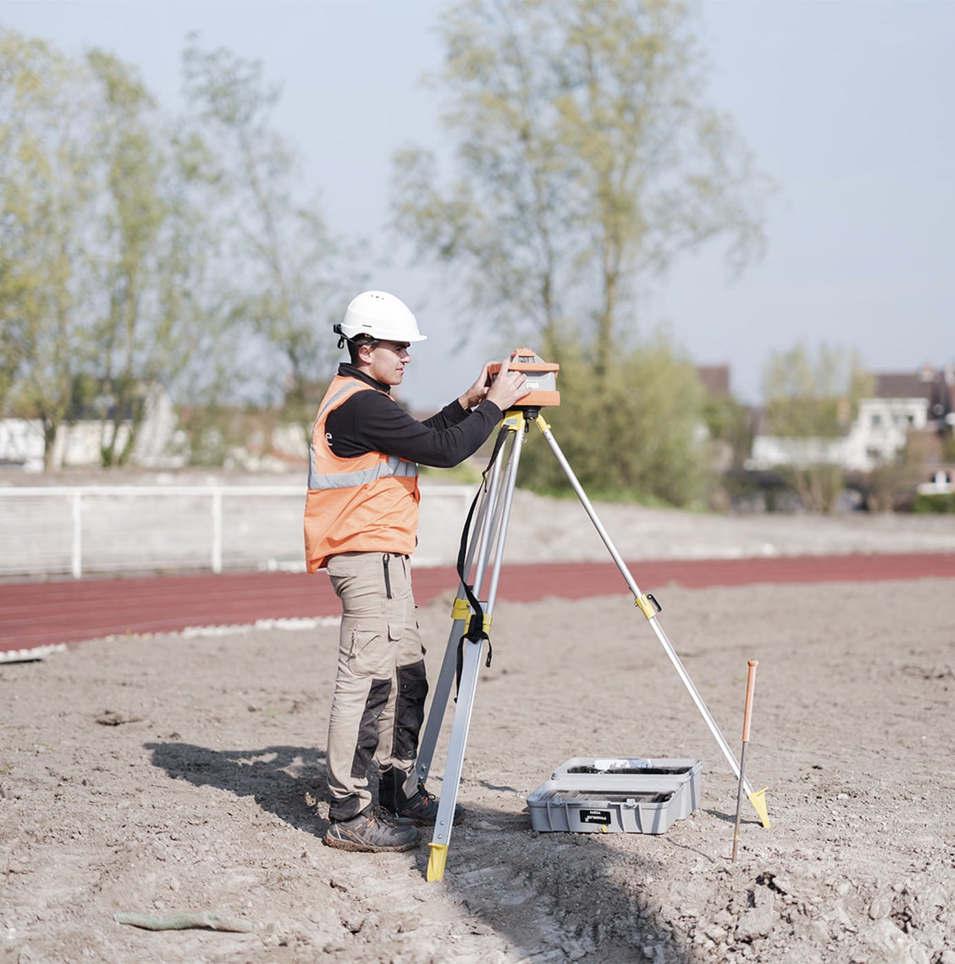Medewerker met veiligheidshelm en oranje hesje verricht landmeting met een theodoliet op een kaal terrein bij een sportveld.