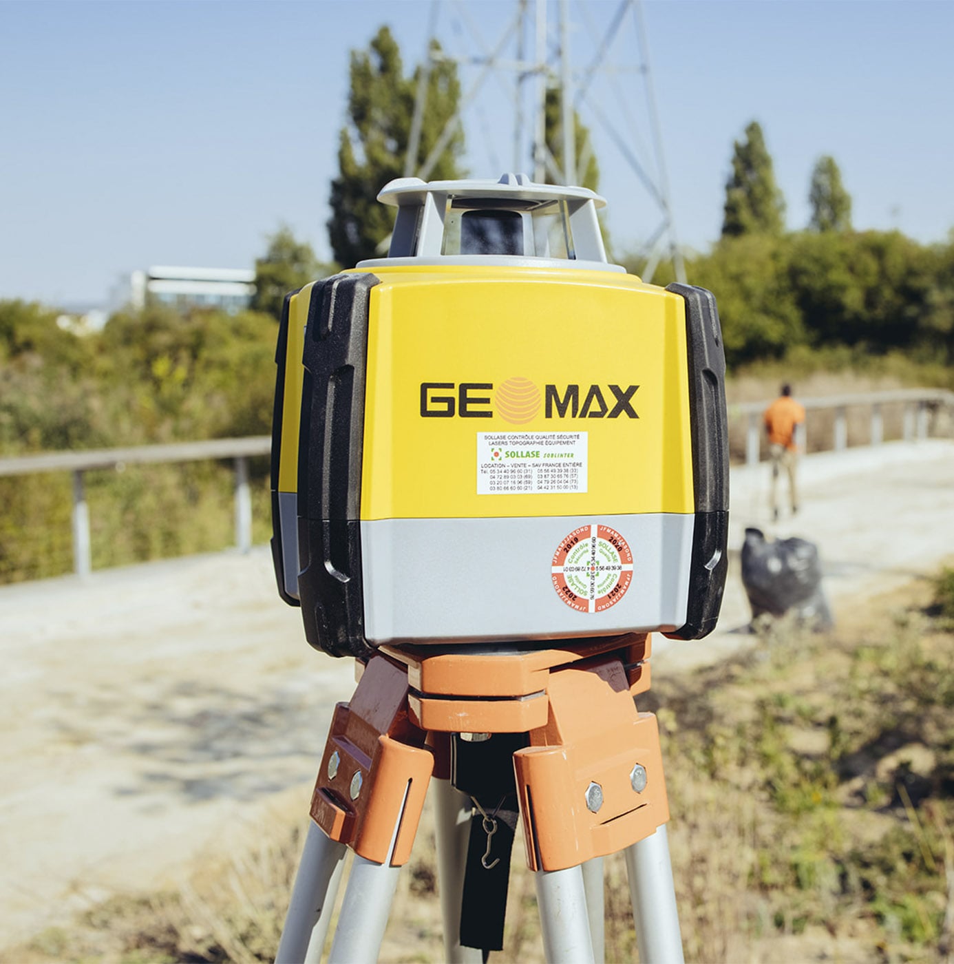 Rotatielaser op statief voor landmeetkundig werk op een buitenlocatie, met een medewerker op de achtergrond.
