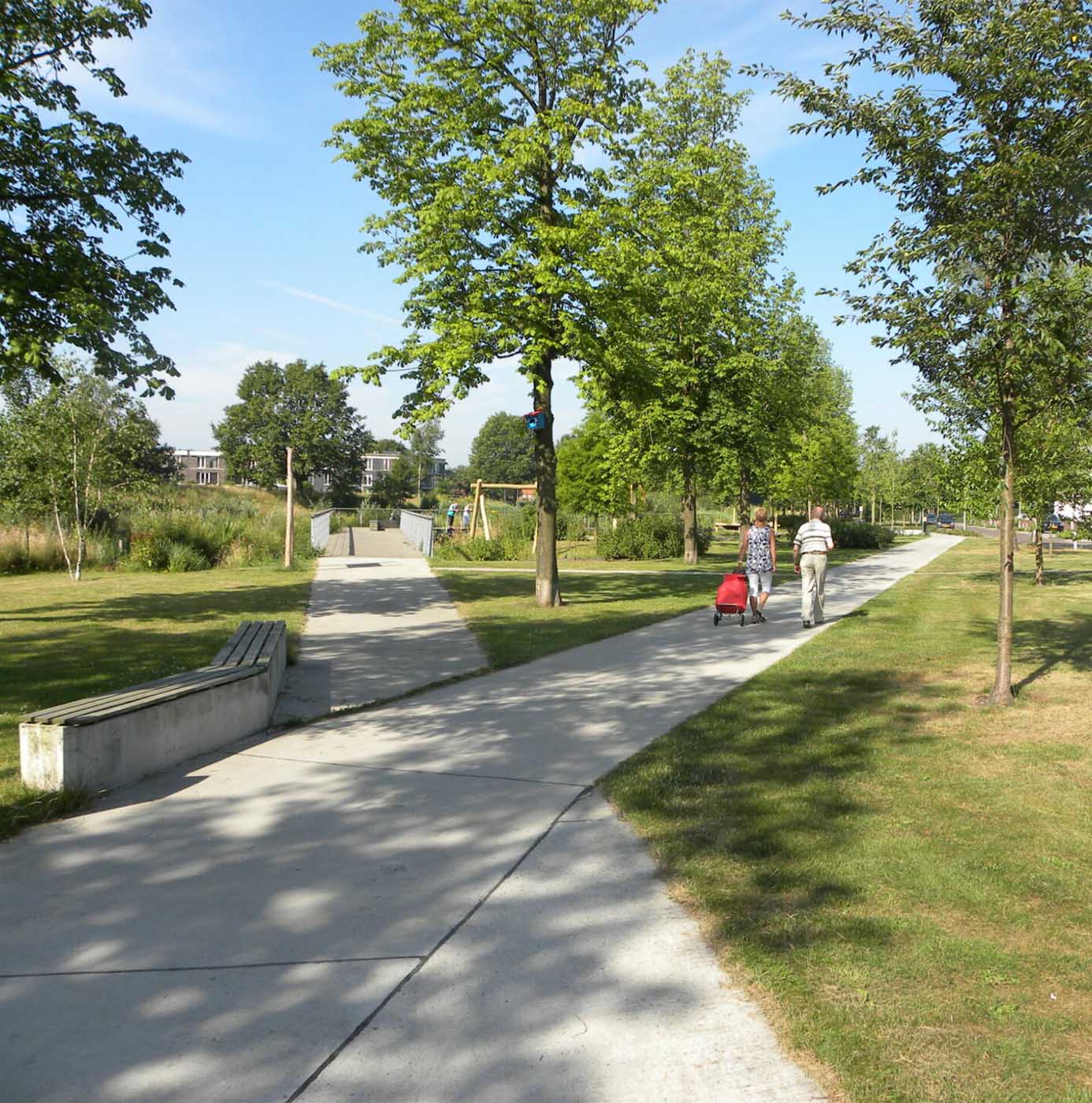 Wandelaars op verhard pad door groen ingericht park met gazon, jonge bomen en bankjes.