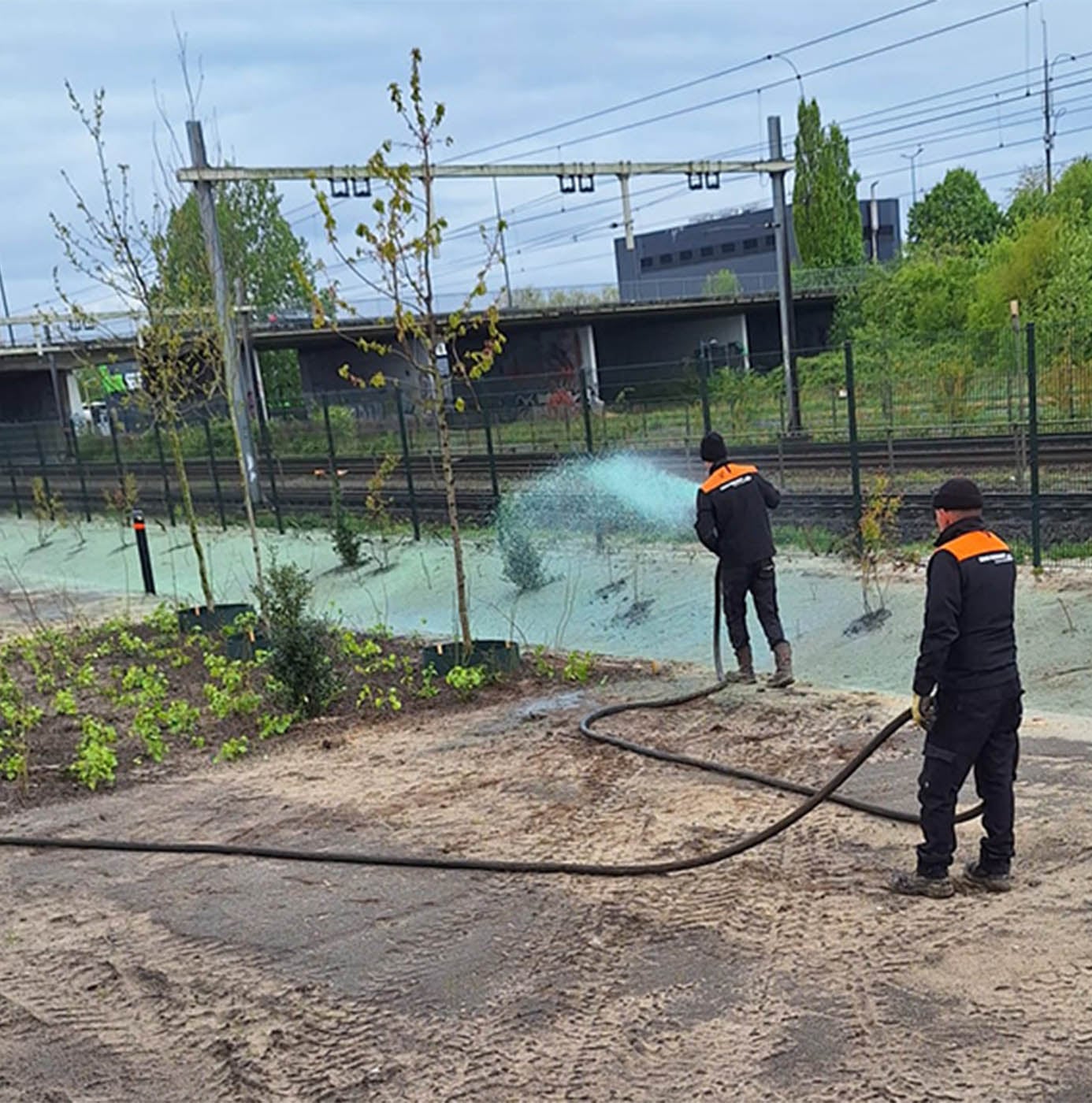 Medewerkers brengen met een slang hydromulch aan op een pas beplante grondstrook naast een spoorbaan.