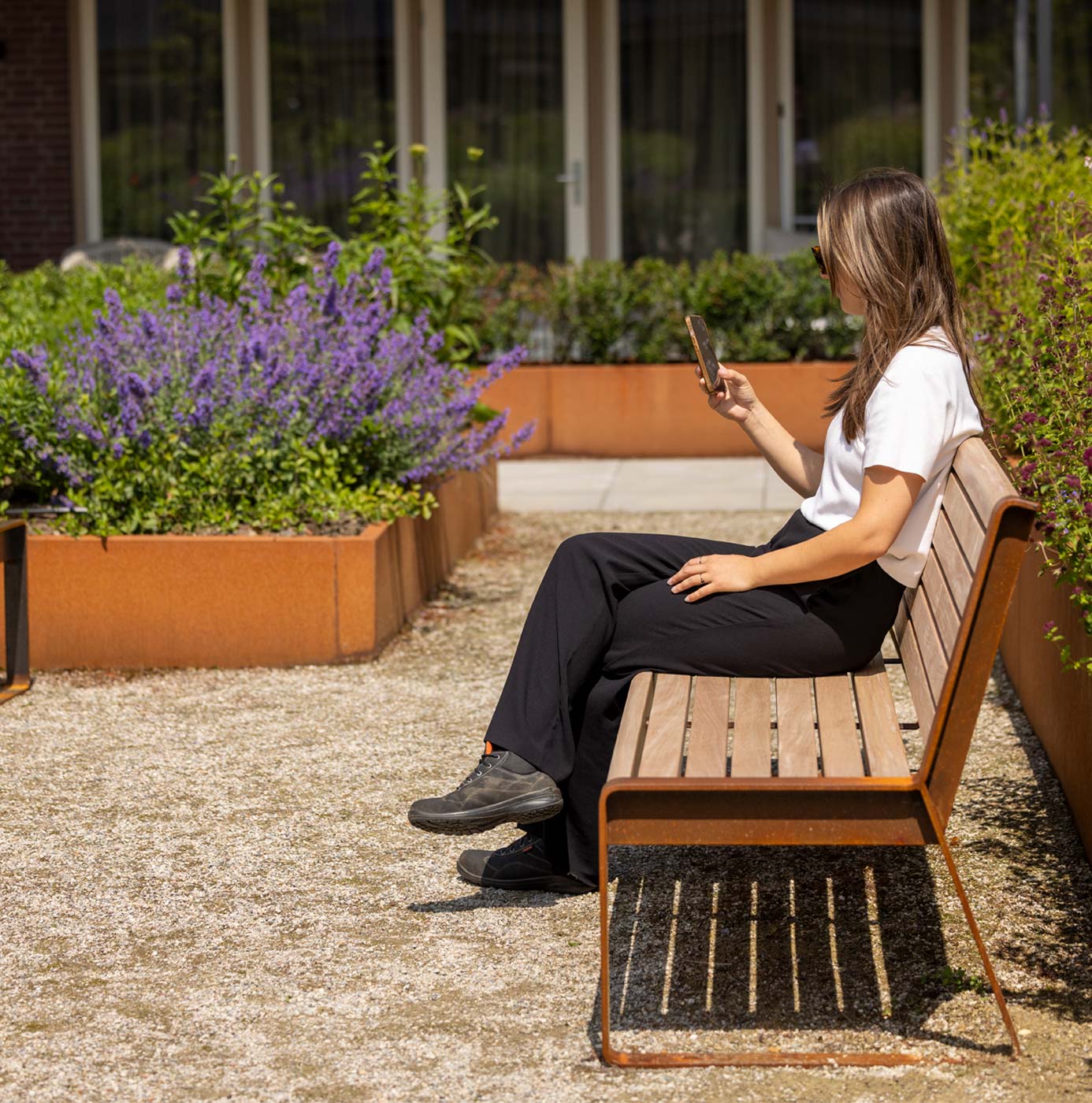 Vrouw zit op houten bankje bij plantenbakken met lavendel tussen verhoogde cortenstalen border beplanting.