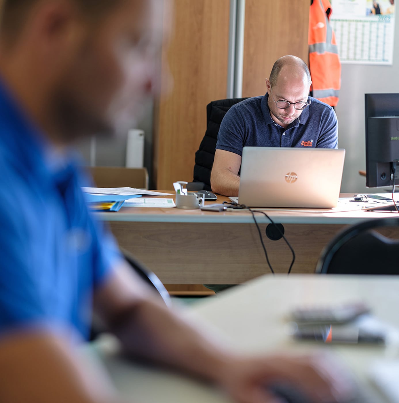 Medewerkers werken op kantoor achter laptops, met een veiligheidshesje op de achtergrond.
