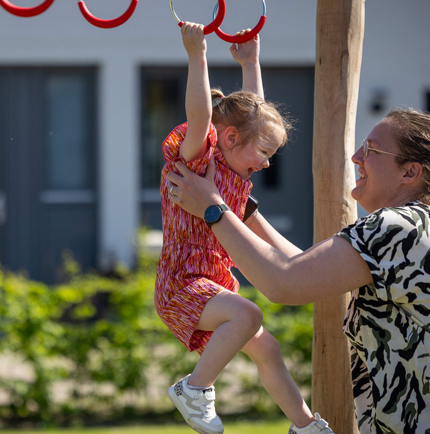 Kind hangt aan ringen van speeltoestel in woonwijk, begeleid door volwassene.