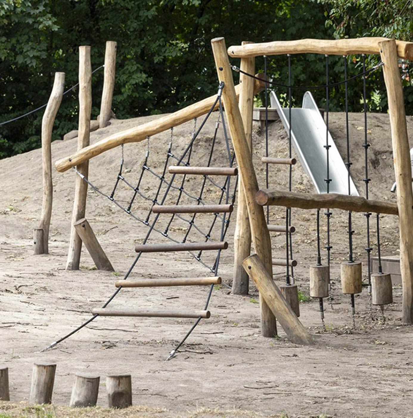 Natuurlijk speeltoestel van robuuste boomstammen met klimnet, glijbaan en balanceerpalen op een zandheuvel.