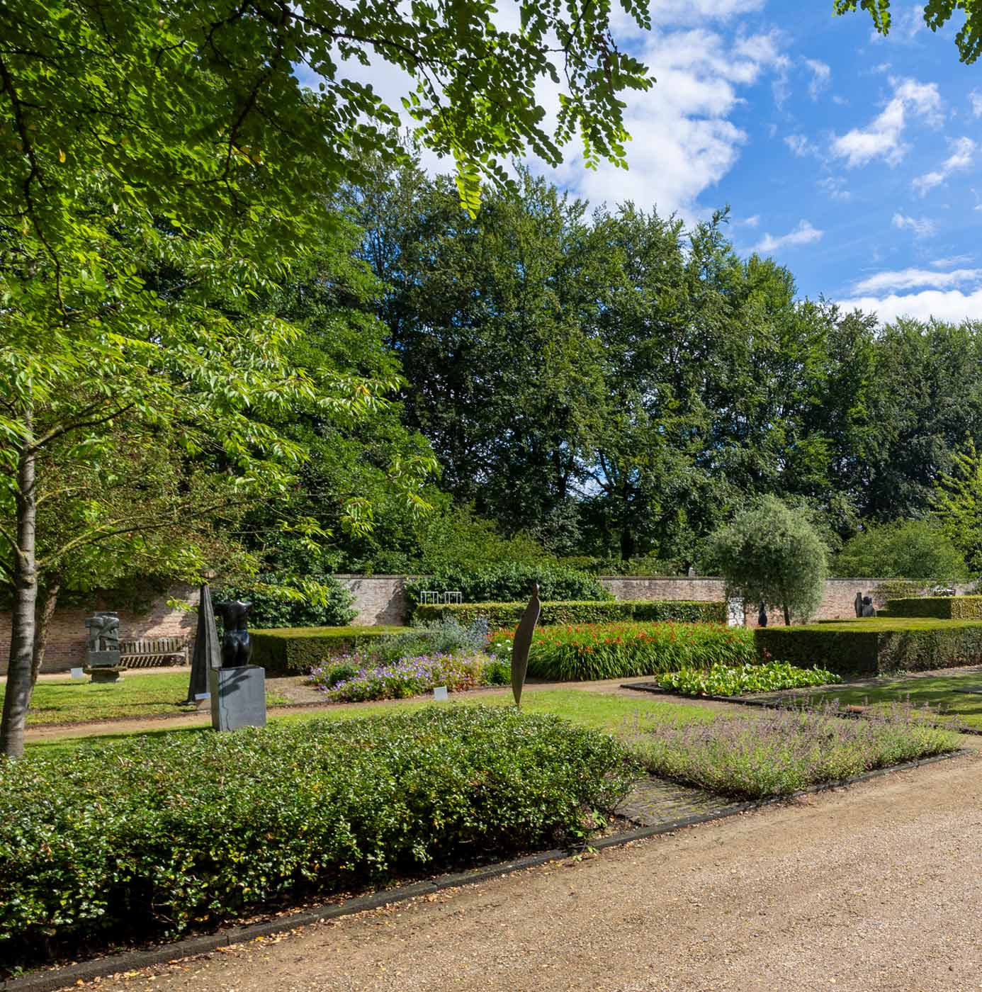 Aangelegde tuin met strak gesnoeide hagen, bloemperken en beeldenkunst omringd door bomen.