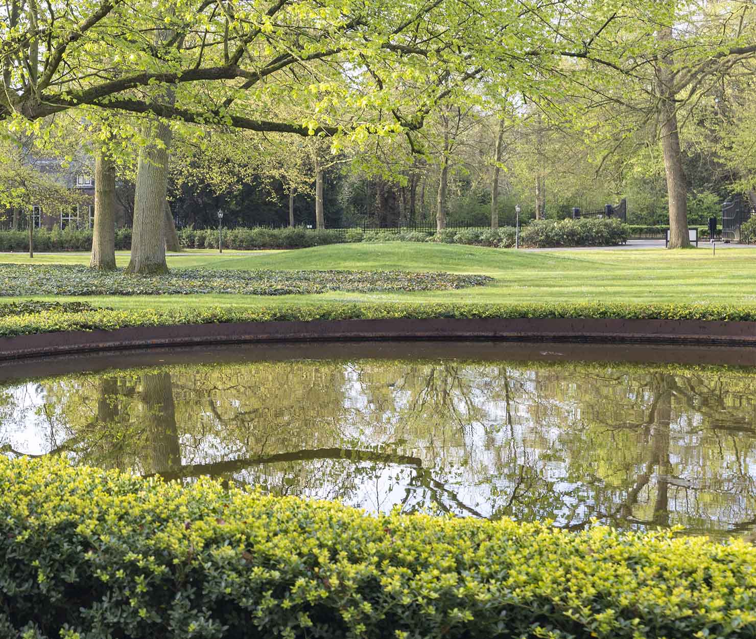 Formele parktuin met vijver, gemillimeterd gazon en strak gesnoeide hagen omgeven door bomen.
