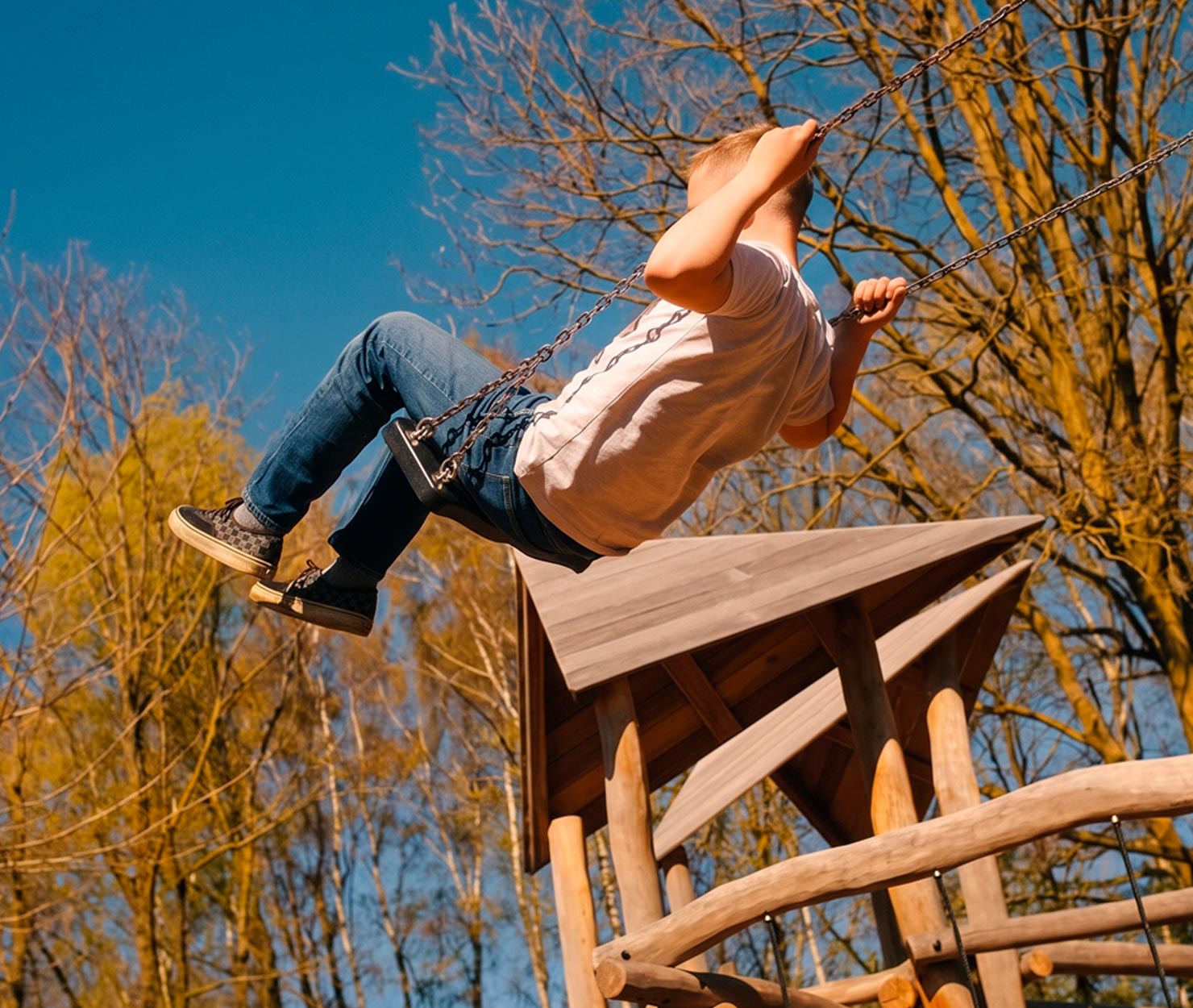 Kind schommelt hoog op speeltoestel bij een houten speelhuisje in een groene speeltuin.
