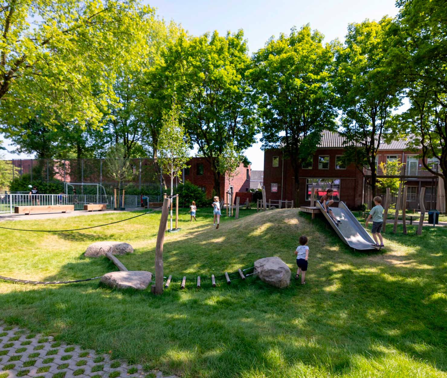 Kinderen spelen op een natuurlijke speelheuvel met glijbaan, balanceerbalken en klimbomen in een groene woonwijk.