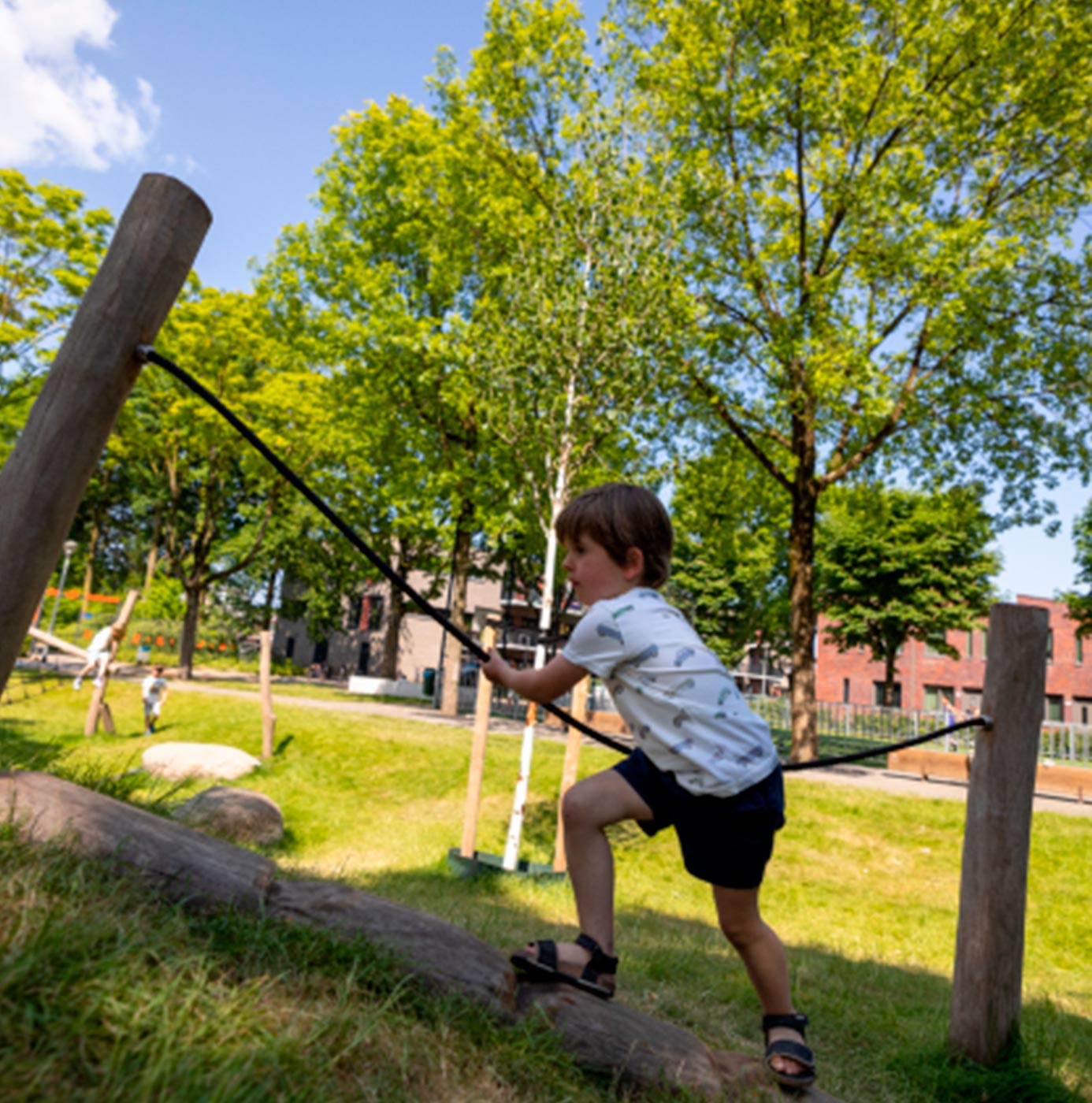 Kind klimt over boomstam met touw op natuurlijke speelplek in groene woonwijk.