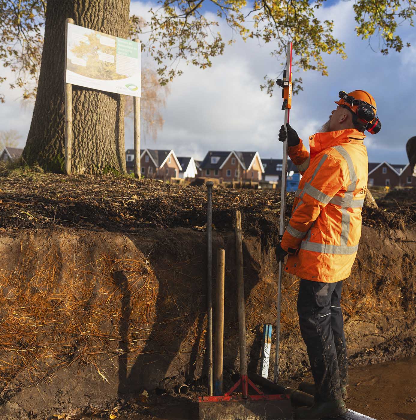 Medewerker met veiligheidshelm en gehoorbescherming voert bodemonderzoek uit bij boomwortels met een meetlat naast een bouwlocatie.