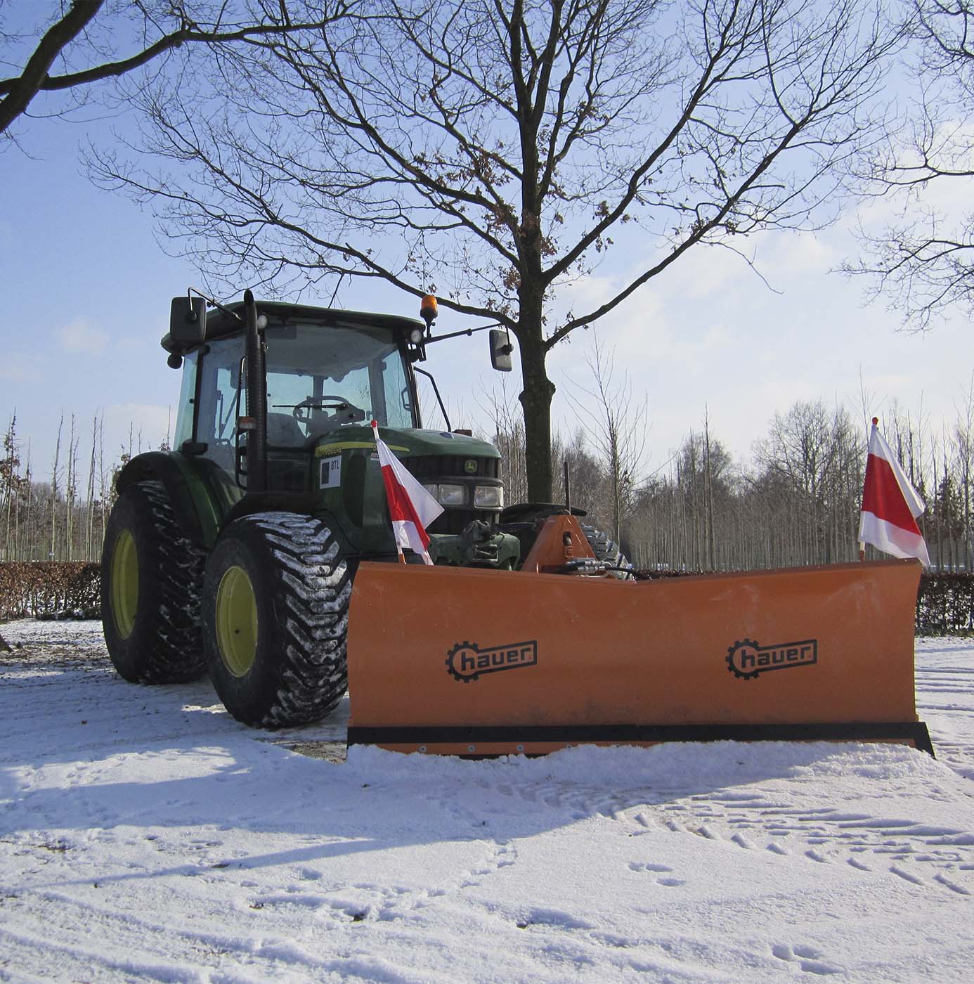 Tractor met sneeuwschuif ingezet voor gladheidsbestrijding op besneeuwd terrein tussen kale bomen.