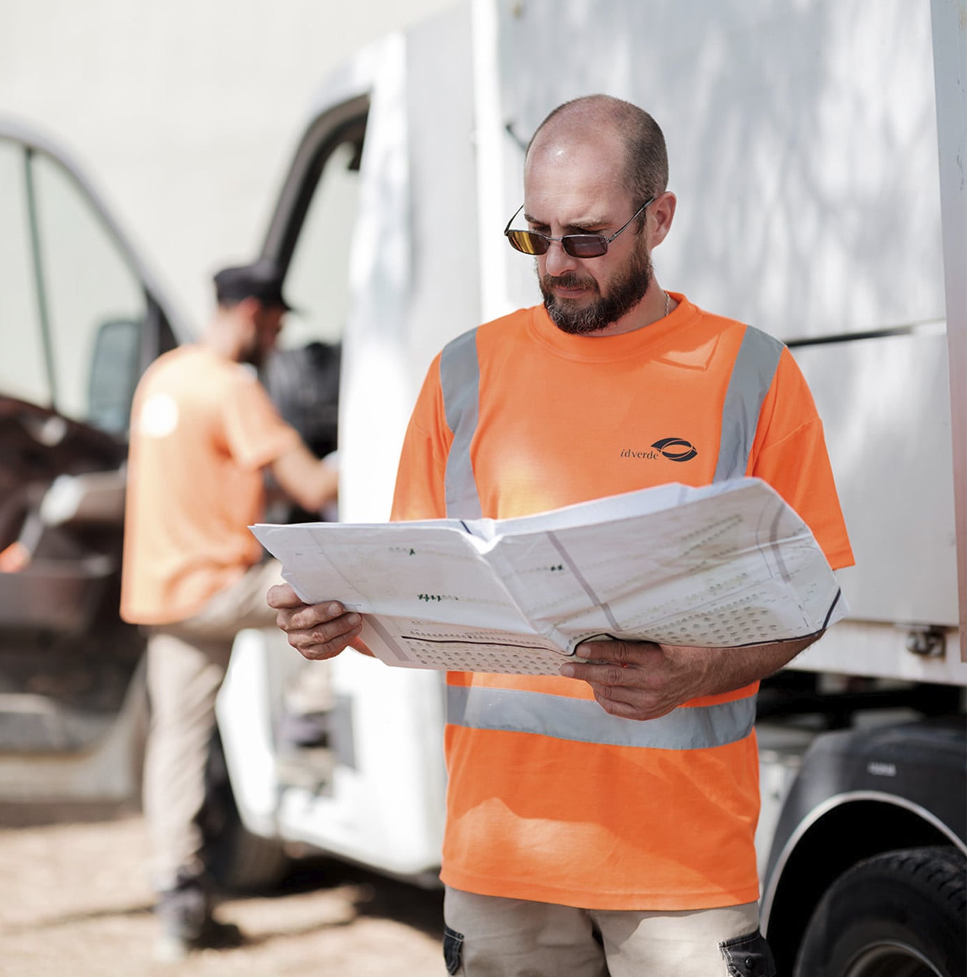 Medewerker in idverde-werkkleding bekijkt werkplanning bij bedrijfsbusje, collega laadt materiaal in de achtergrond.