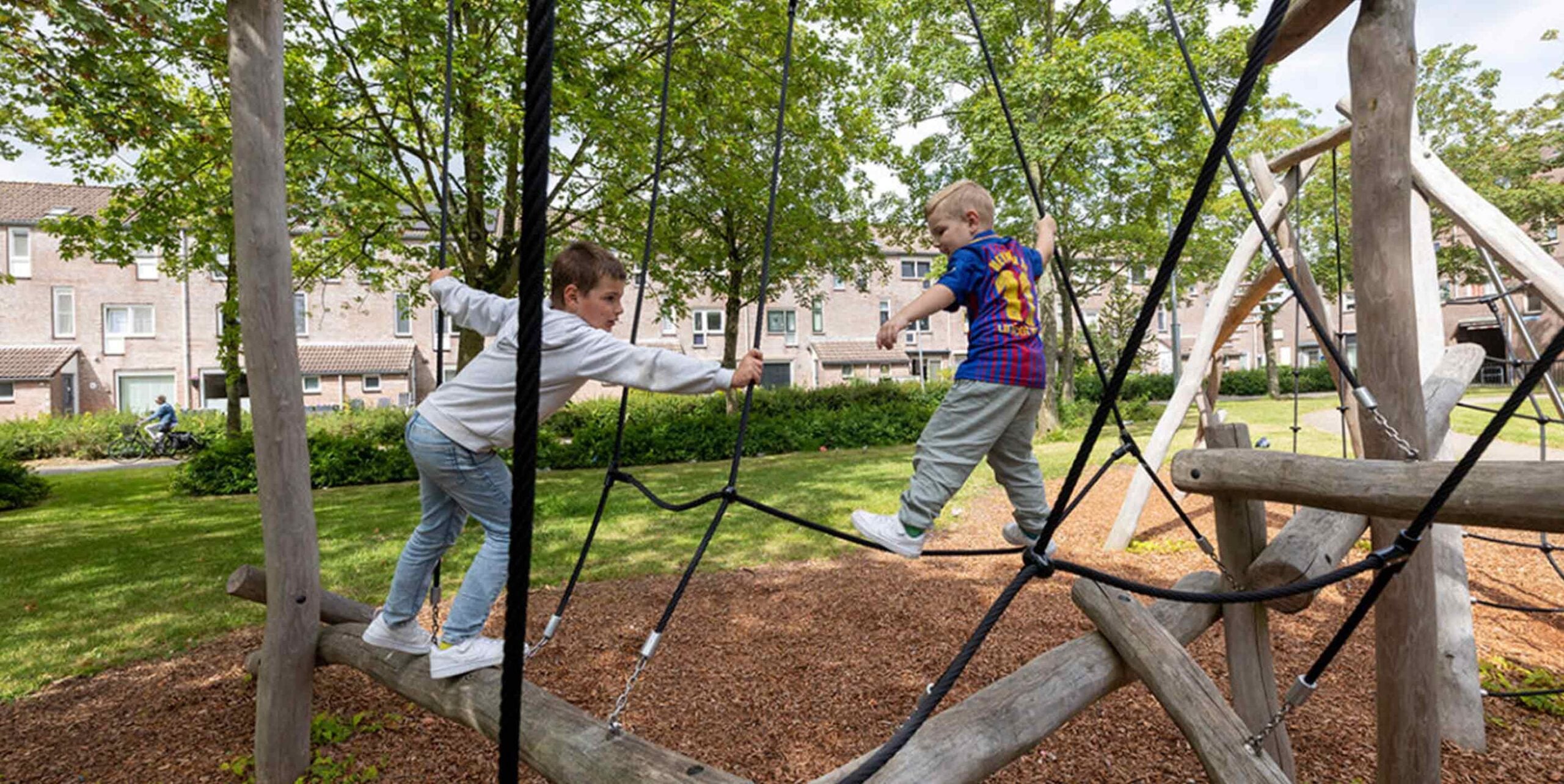 Kinderen klimmen op een houten speeltoestel met touwen in een groene woonwijk.