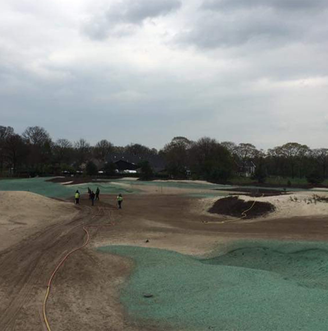 Medewerkers in veiligheidskleding lopen over kale zandvlakte tijdens hydroseeding-werkzaamheden op een golfterrein.