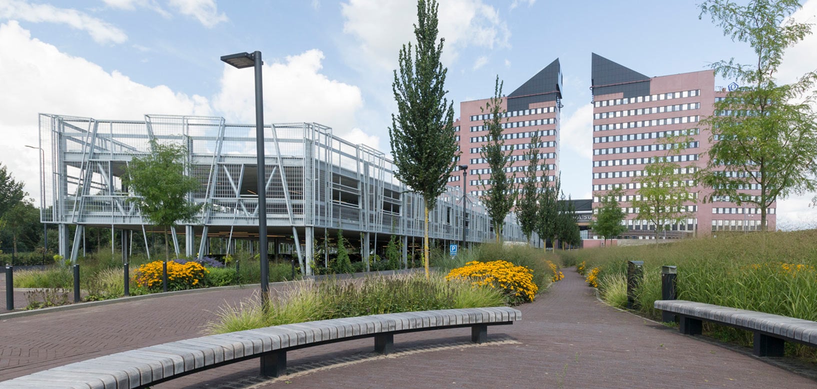 Beplant wandelpad met siergrassen en bloeiende zonnehoed bij kantoorgebouwen en een parkeergarage.