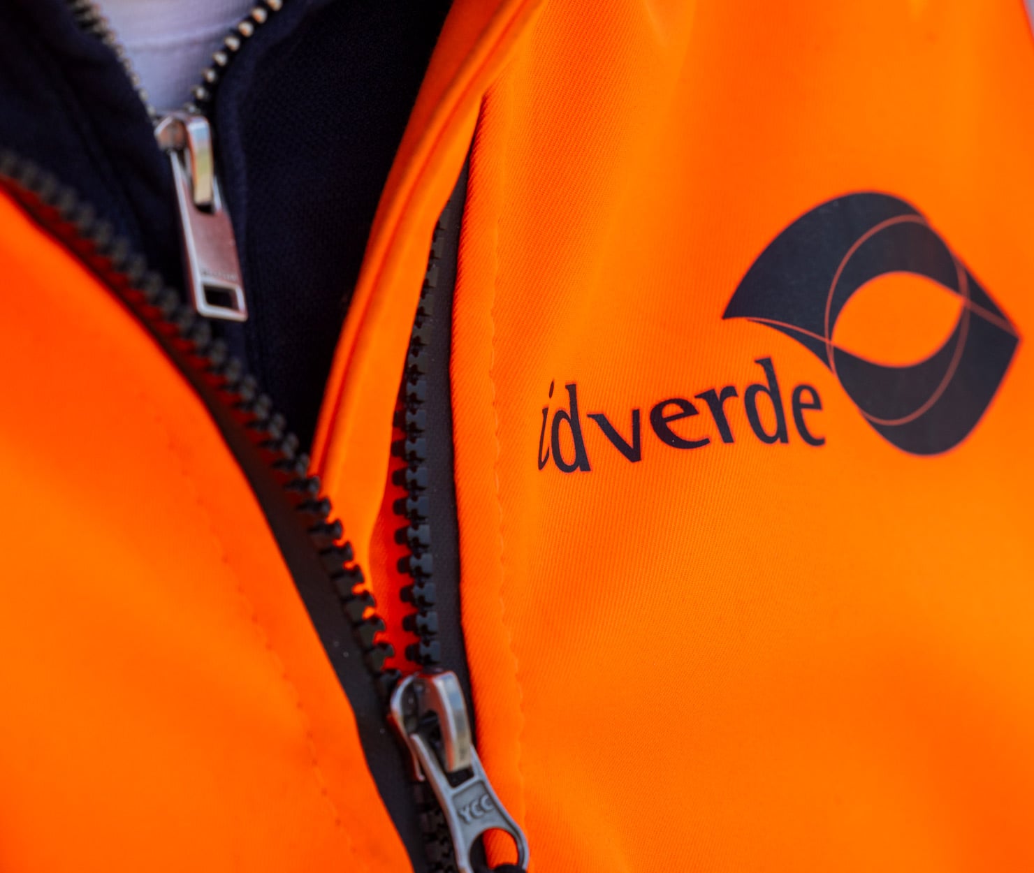 Close-up van oranje werkjas met idverde-logo op de borst.