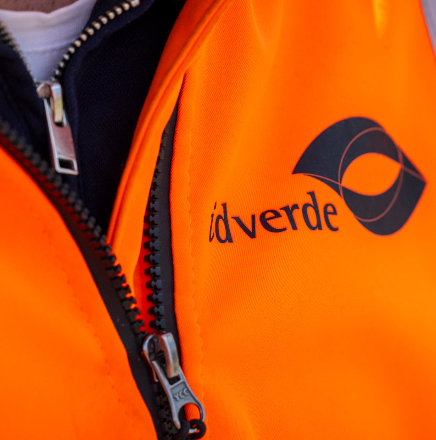 Close-up van oranje werkjas met idverde-logo op de borst.