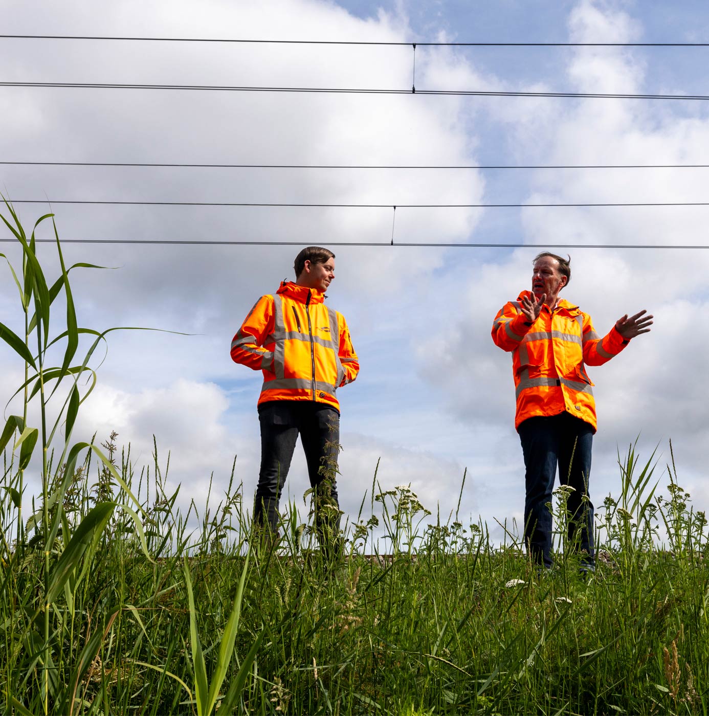 Twee medewerkers in oranje veiligheidsjassen overleggen bij begroeide spoorberm onder bovenleiding.