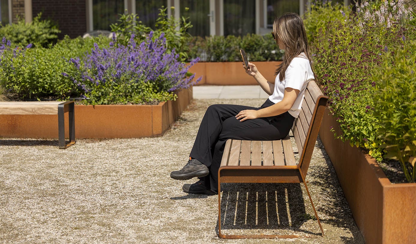 Vrouw zit op een houten bankje tussen cortenstalen plantenbakken met bloeiende beplanting in een bedrijfstuin.