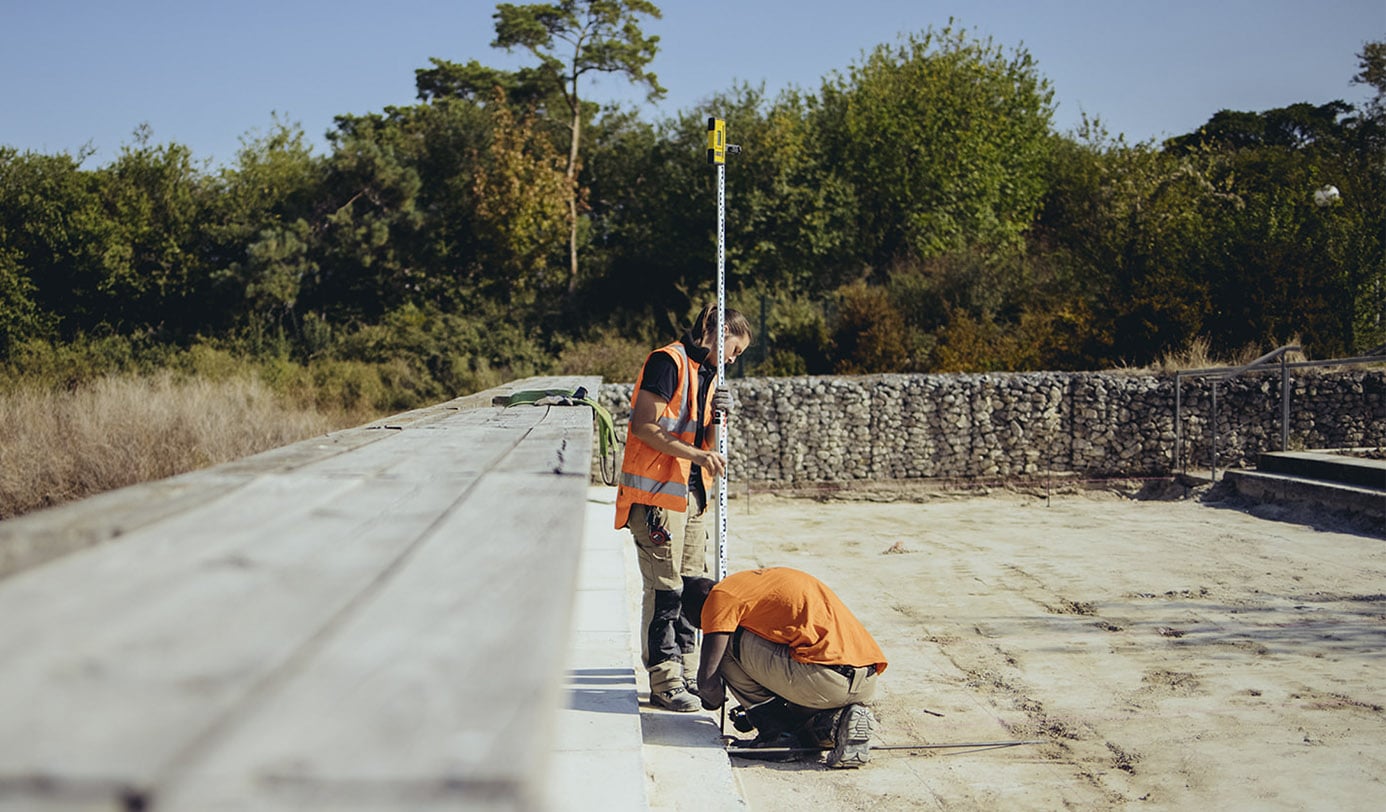 Medewerkers in oranje werkkleding verrichten hoogtemetingen met een meetlat bij een gabionmuur op een zandvlakte.