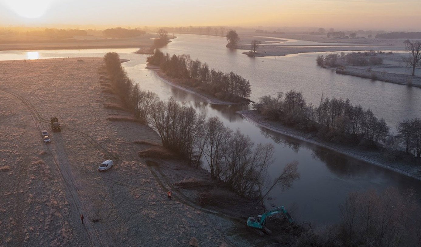 Luchtfoto van rivieroever met graafmachine en werkverkeer tijdens onderhoud aan de uiterwaarden bij zonsopkomst.