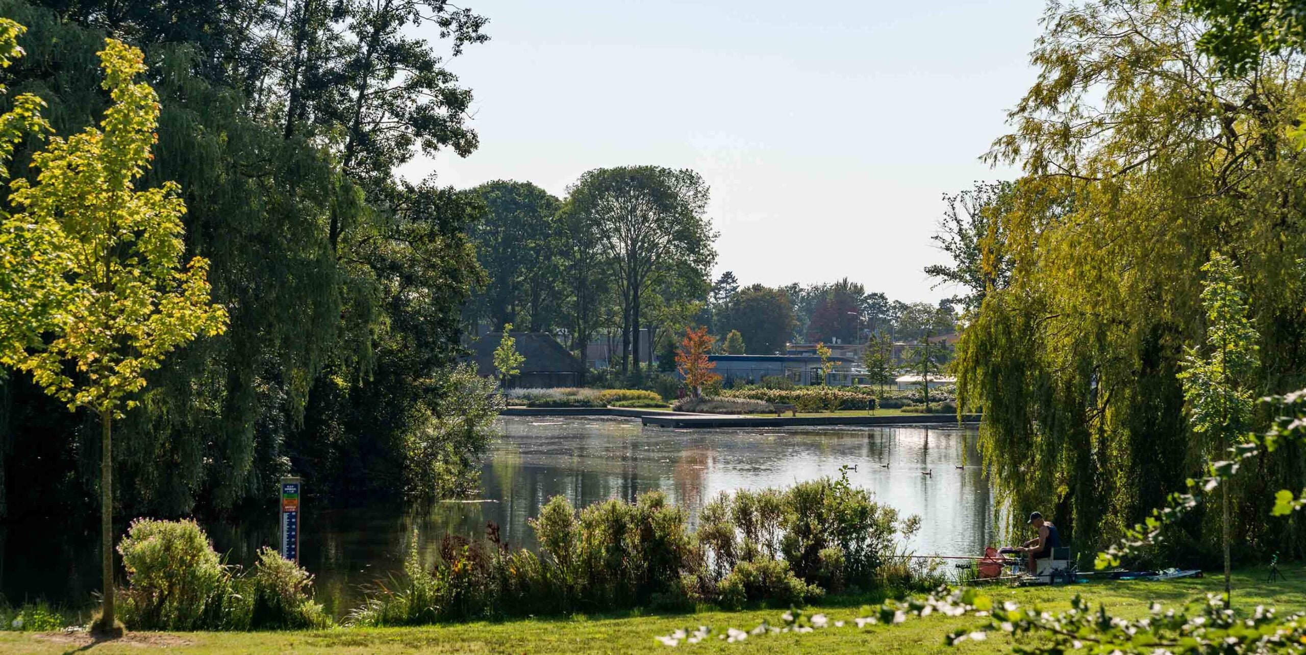 Groen ingericht waterpark met vijver, oevergroen en een visser aan de waterkant.