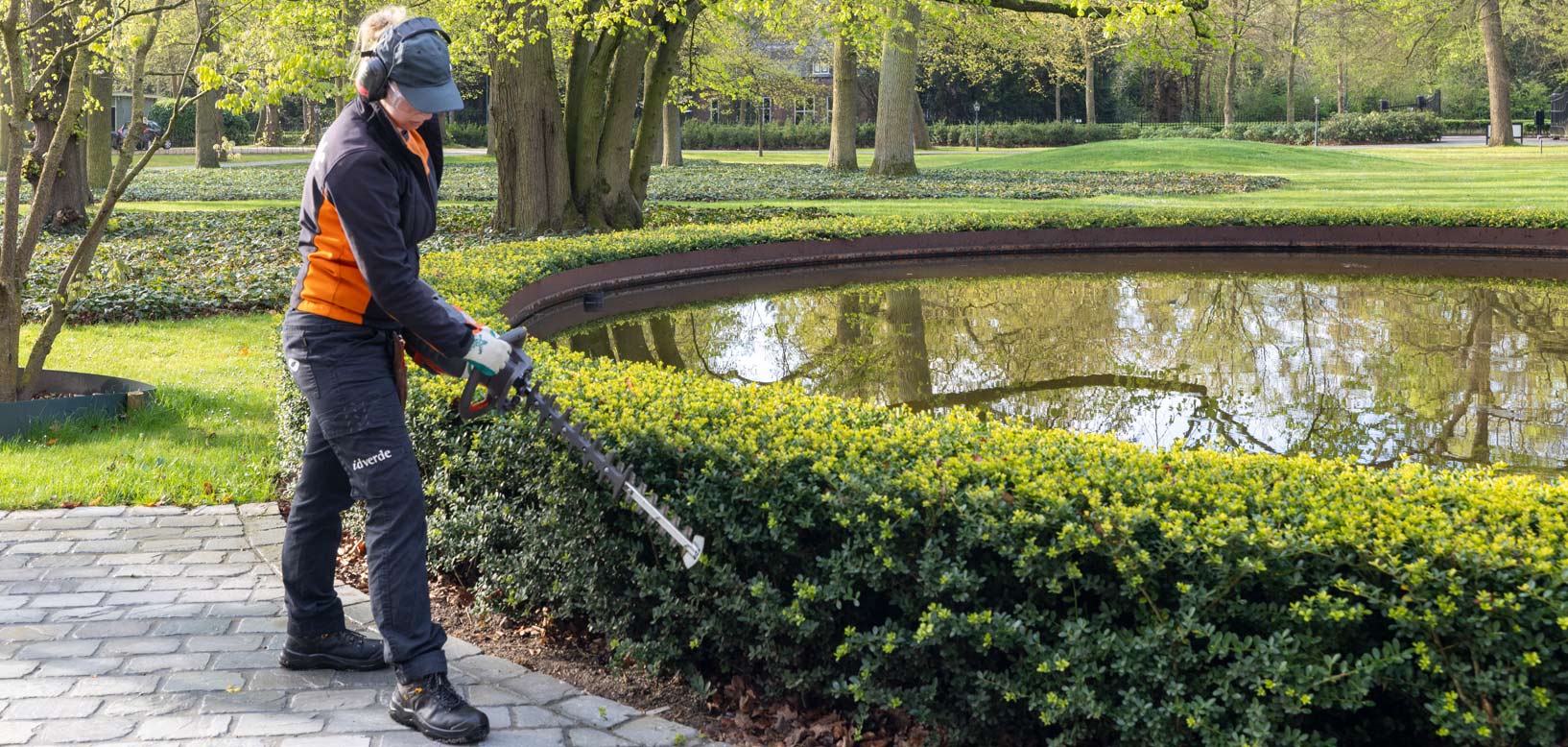 Medewerker van idverde snoeit een lage haag langs een vijver in een park met heggenschaar.