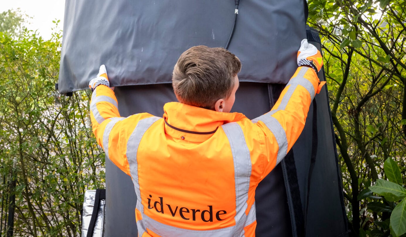 Medewerker van idverde in oranje werkkleding bevestigt een afdekhoes over een verkeersinstallatie tussen struiken.