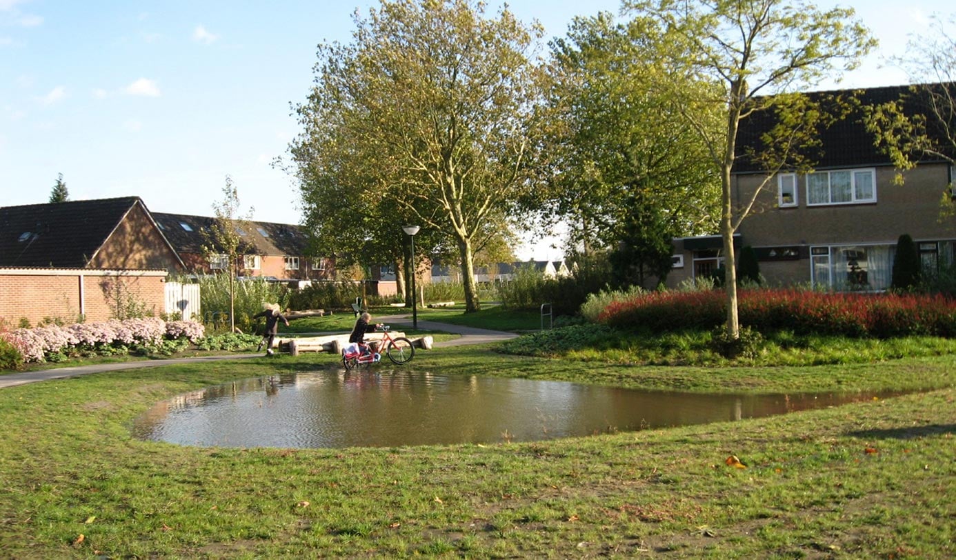 Groene woonwijk met waterpartij, gazon, beplanting en spelende kinderen tussen de woningen.