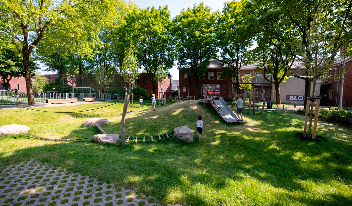Kinderen spelen op een groen ingericht schoolplein met glijbaan, klimbalken en natuurlijke speelelementen tussen bomen.