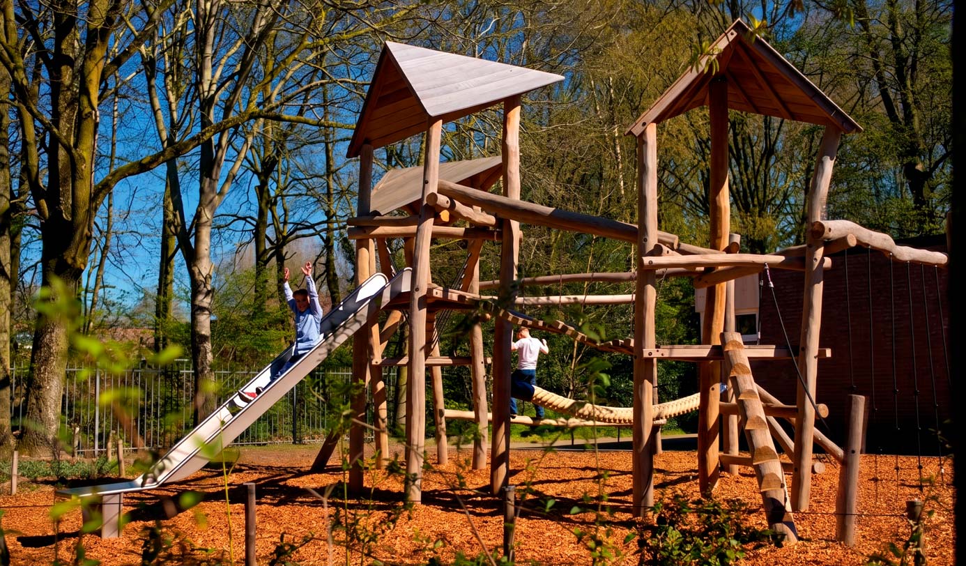 Houten speeltoestel met glijbaan en klimtoren op een houtsnipperondergrond tussen bomen, met spelende kinderen.
