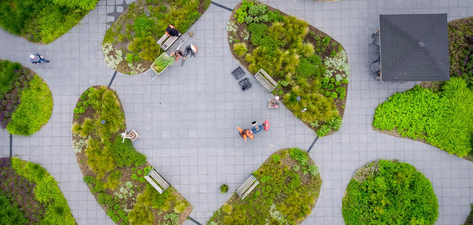 Luchtfoto van medewerkers die groenperken onderhouden op een tegelplein met ronde plantvakken en zitbanken.