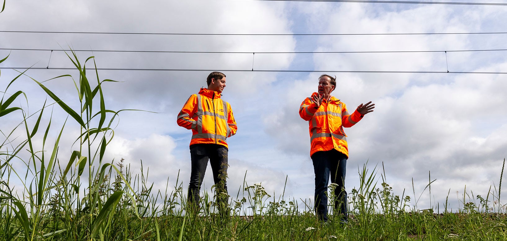 Twee medewerkers in oranje veiligheidsjassen overleggen op een begroeide berm langs het spoor.