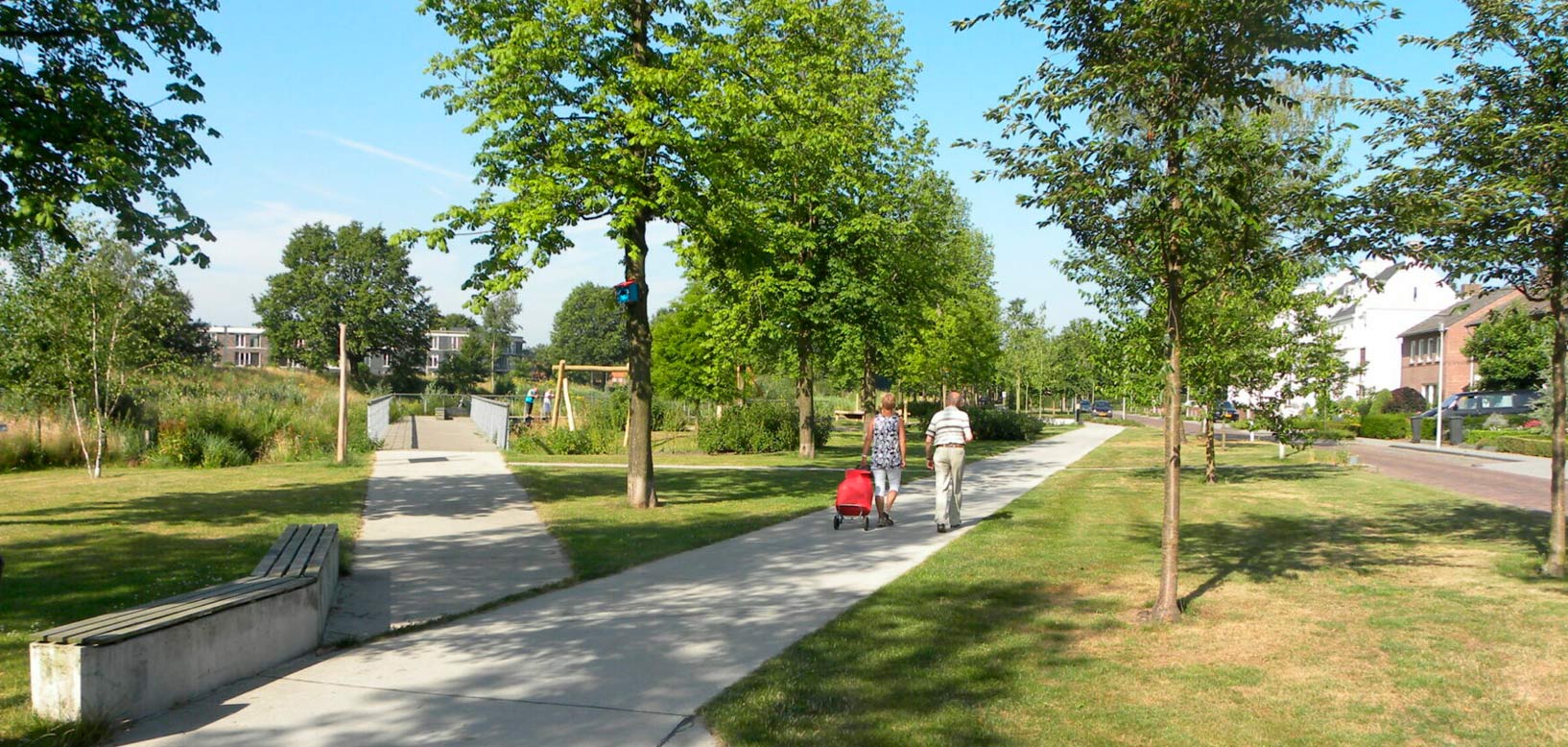 Wandelaars op verhard pad door groen wijkpark met bomenlanen en gazon nabij woonwijk.