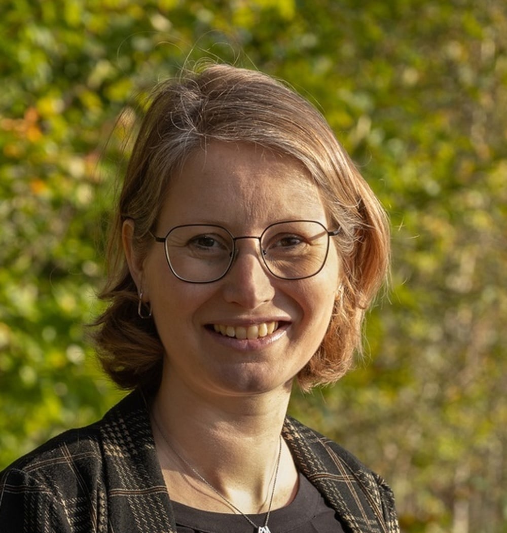 Portret van Ellen van Boxtel, Group Controller bij idverde, met groen gebladerte op de achtergrond.