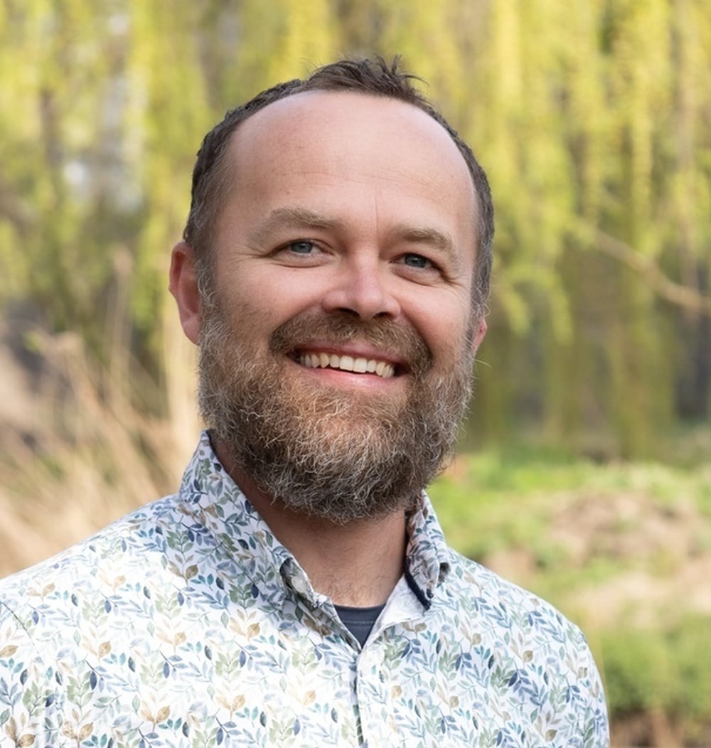 Medewerker van idverde poseert lachend buiten voor een groene achtergrond met wilgentakken.