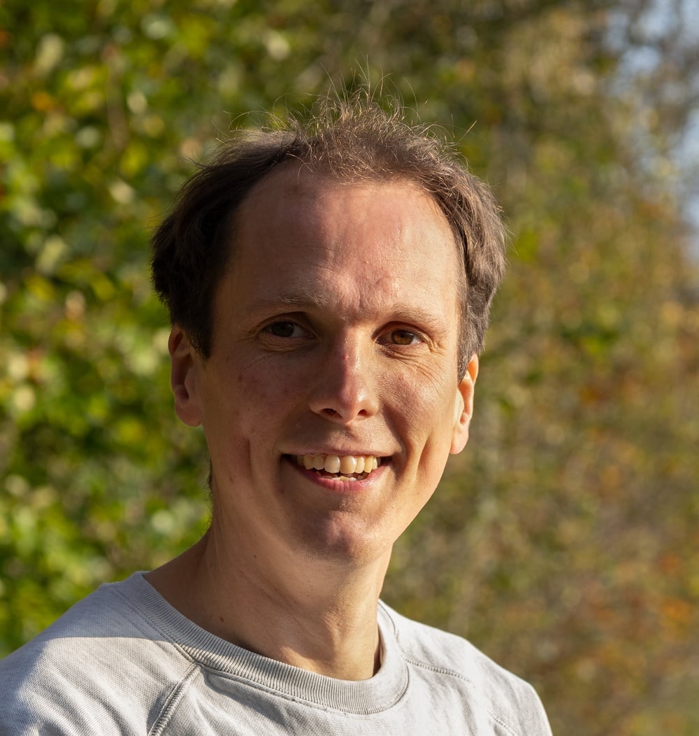Rick Visser, werkvoorbereider Dak- en Gevelgroen, lacht in close-up portret met groene bomen op achtergrond.