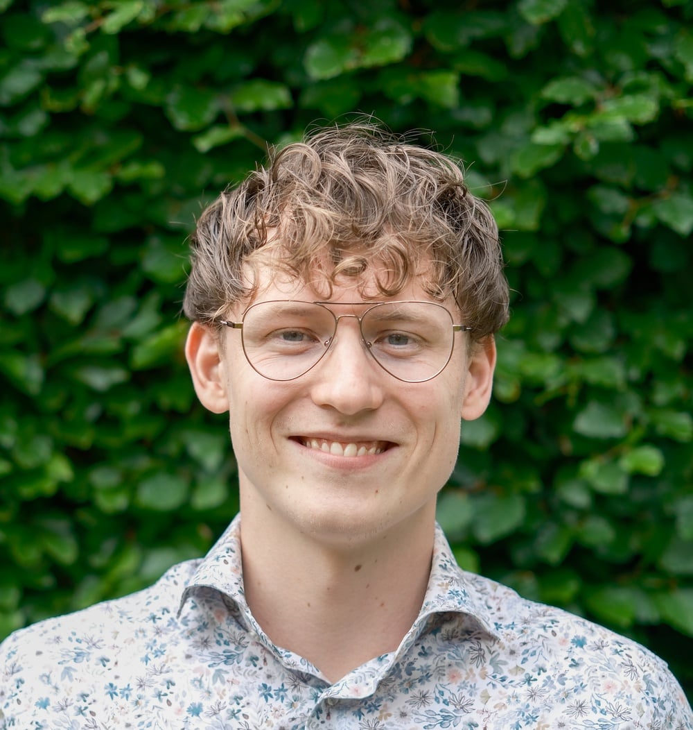 Medewerker met bril poseert lachend voor een groene heg.