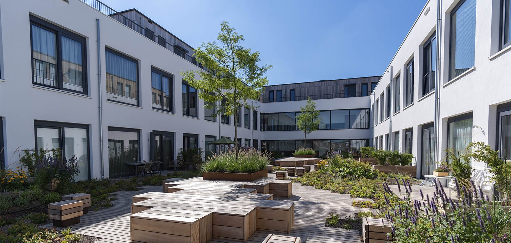 Groene binnentuin met houten terrasdek, plantenborders en bomen tussen appartementengebouwen.