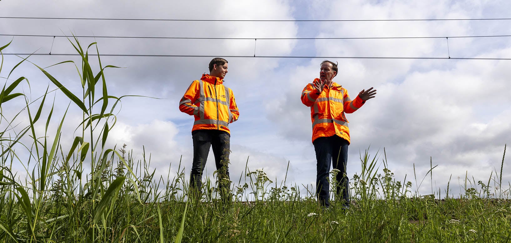 Twee medewerkers in oranje veiligheidsjassen overleggen op een begroeide spoortalud onder bovenleidingdraden.